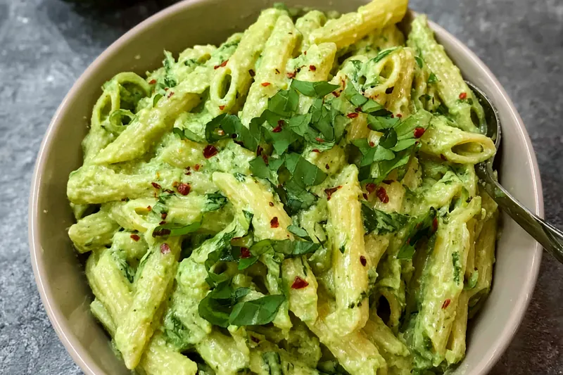 Ingredients for avocado pasta including ripe avocados, fresh basil, garlic, lemon, pasta, and cherry tomatoes arranged on a wooden cutting board